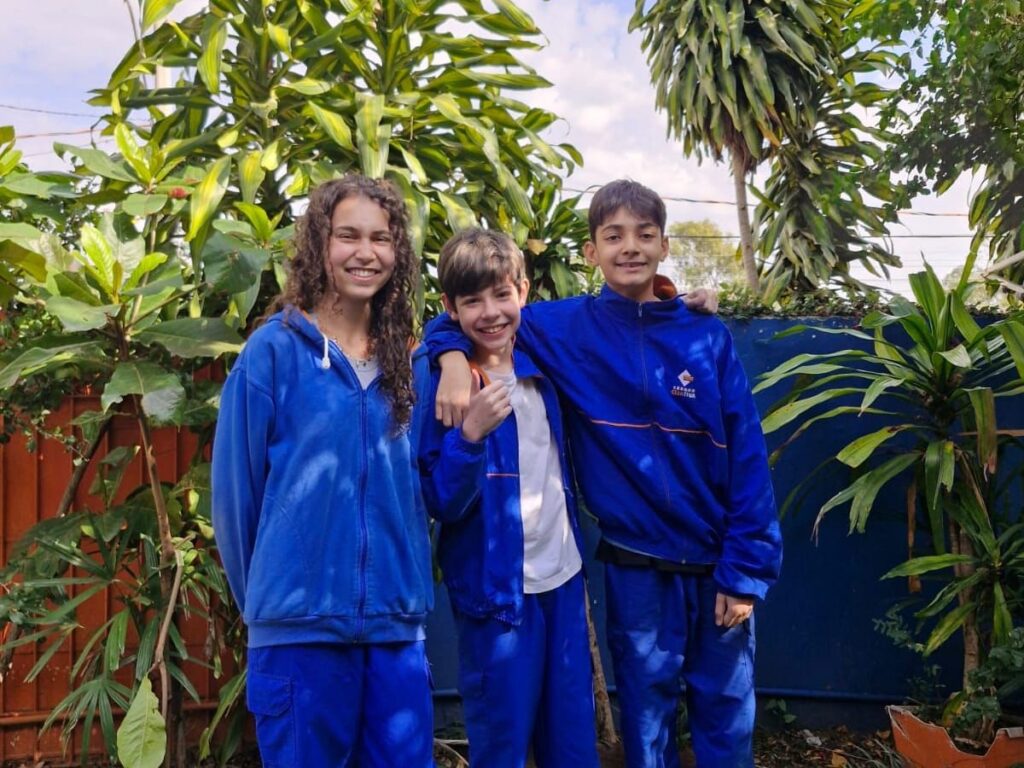 Três alunos do 8º ano, com o uniforme azul da escola, sorriem lado a lado em um jardim.