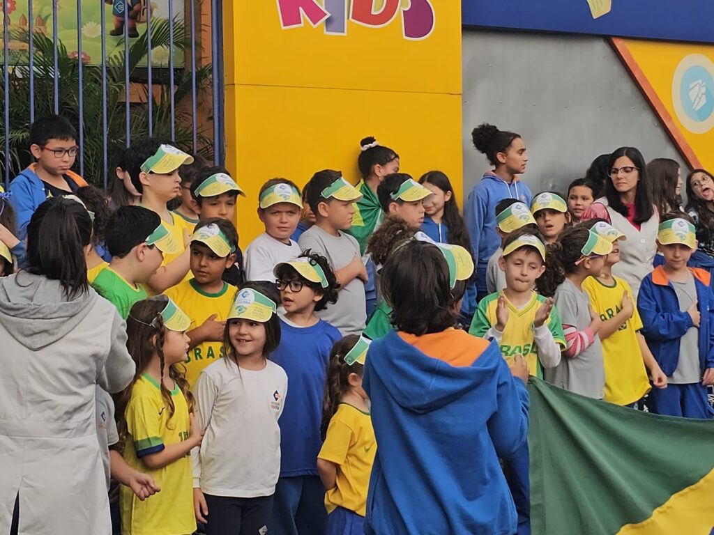 Alunos de uniforme e boné reunidos em frente à escola, com a bandeira do Brasil, na caminhada de 7 de Setembro.