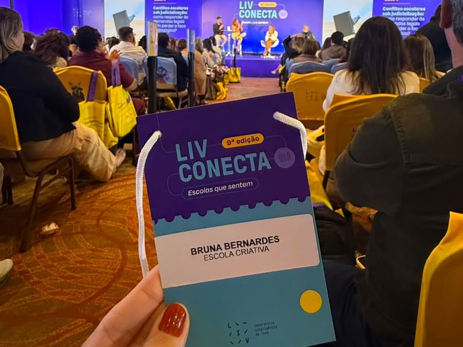 Crachá do LIV Conecta 2026 em primeiro plano, com o auditório do evento ao fundo.